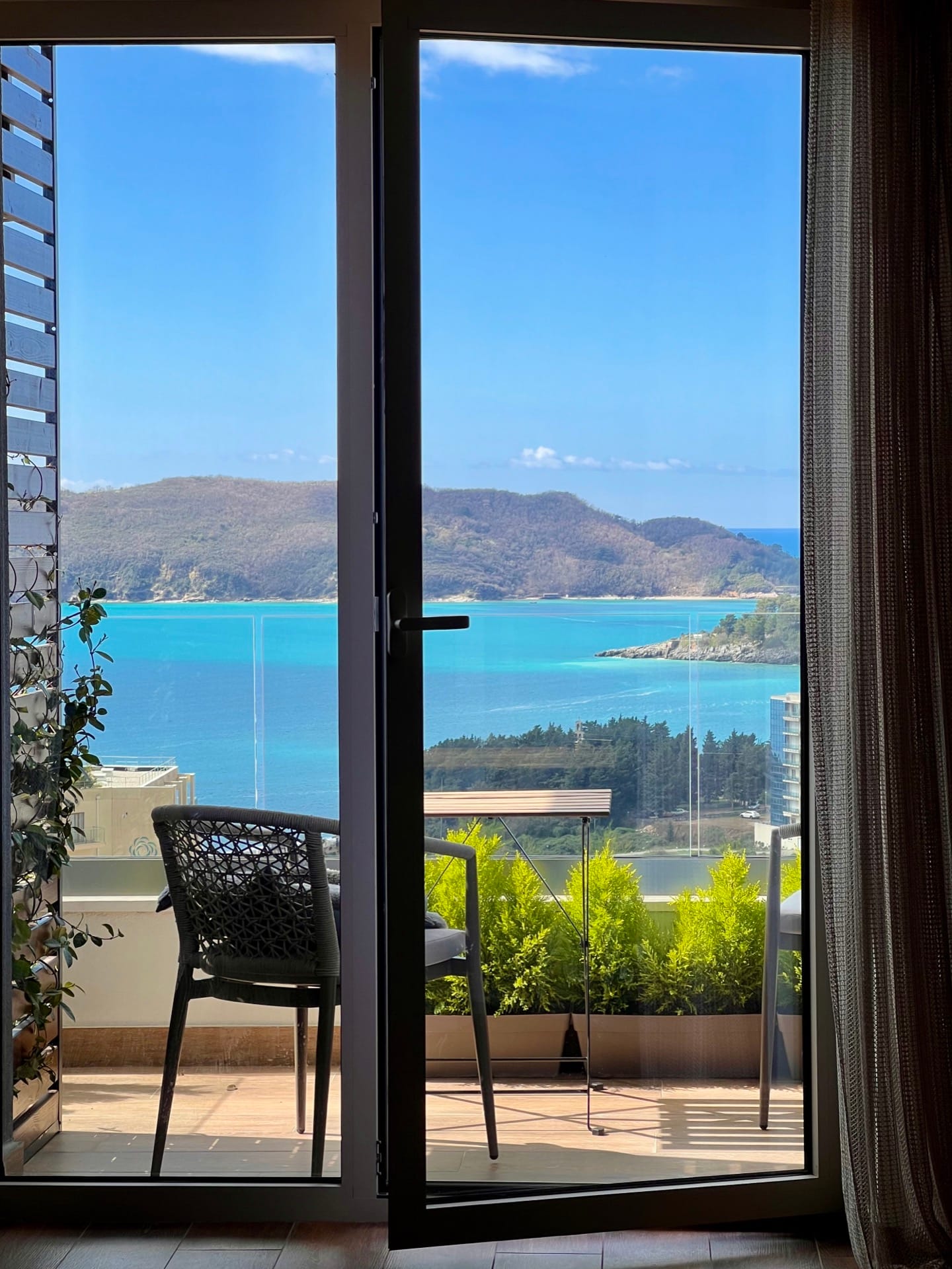 View from the apartment balcony in Bečići, Montenegro: wicker furniture, green plants, and a panoramic view of the Adriatic Sea.