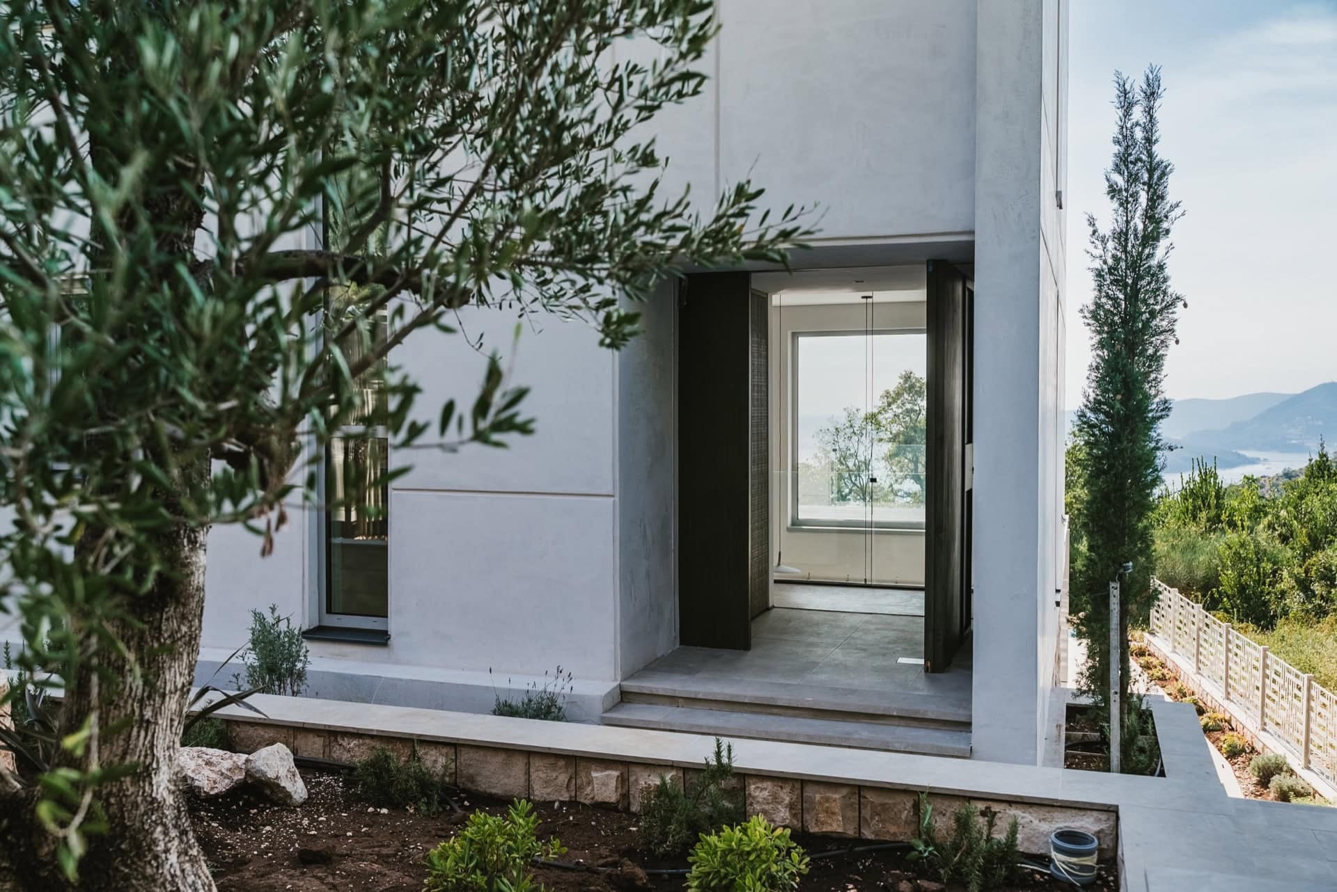 Modern villa in Blizikuće, Montenegro: view of the house entrance with panoramic glazing and a terrace among olive trees.