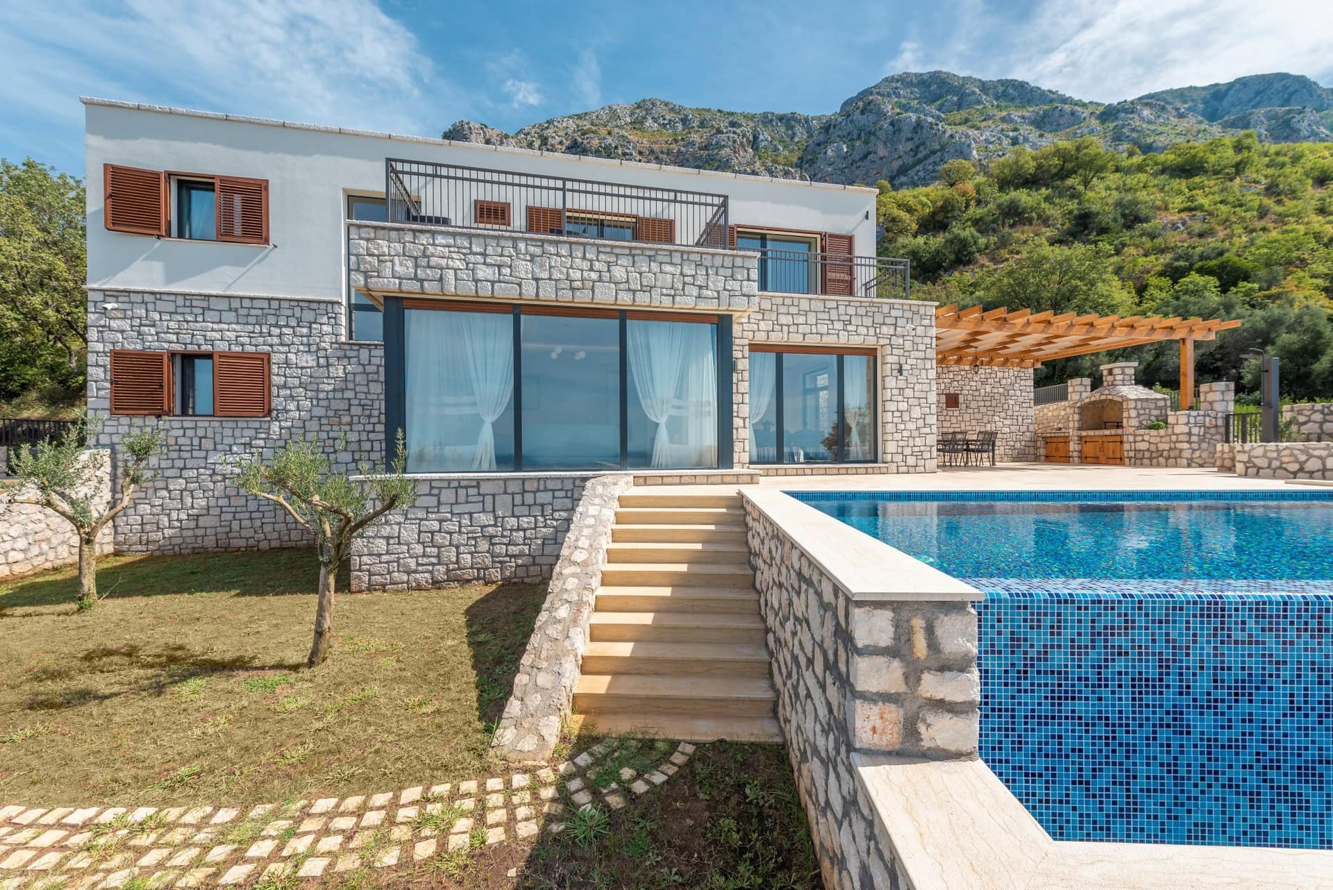 Modern villa in Reževići, Montenegro: stone cladding, panoramic windows, pool, and barbecue area against a mountain backdrop.
