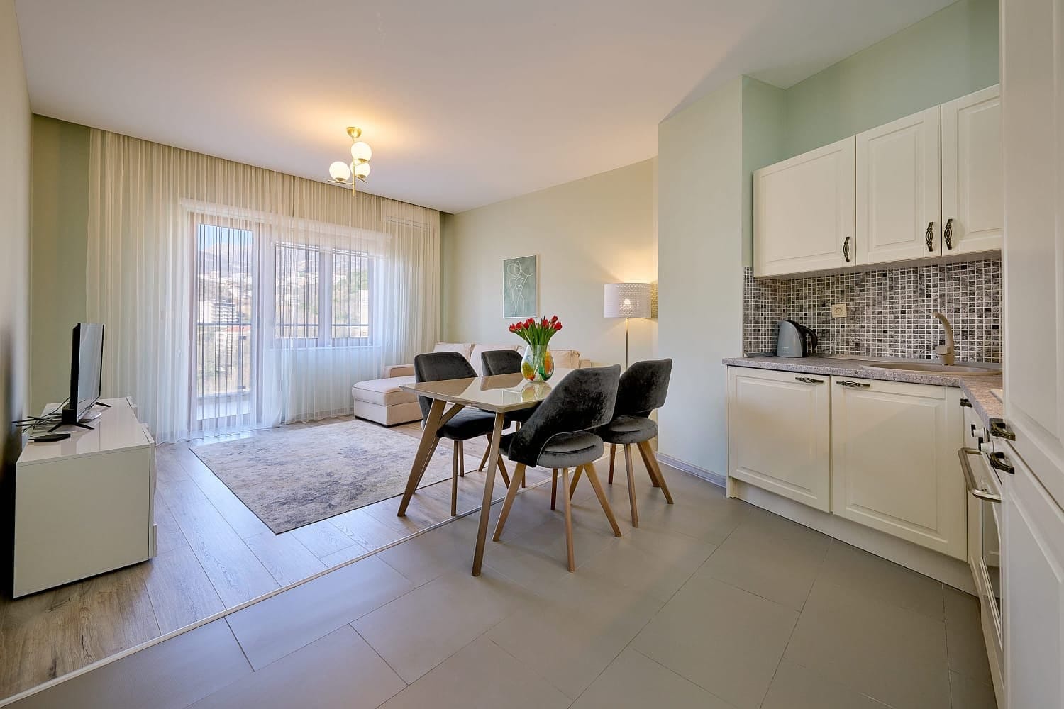 Living room with kitchen in an apartment in Becici, Montenegro: light-colored furniture, dining area, balcony access, and panoramic windows.