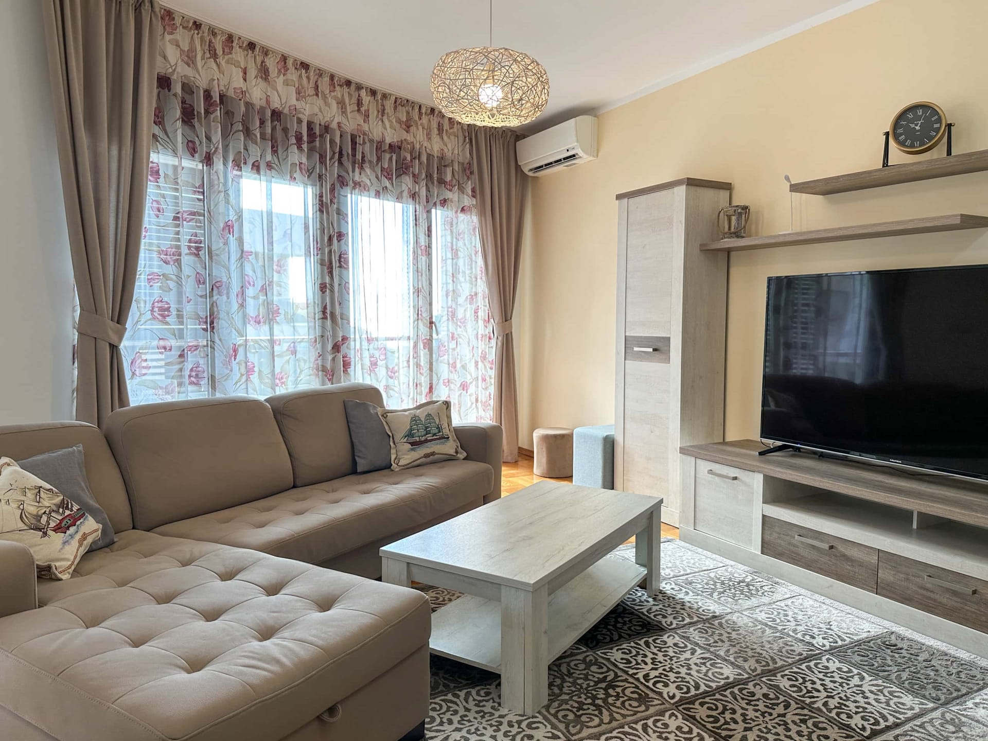 Cozy living room in an apartment in Bečići, Montenegro: sectional sofa, TV area, light-colored furniture, and a panoramic window with sheer curtains.