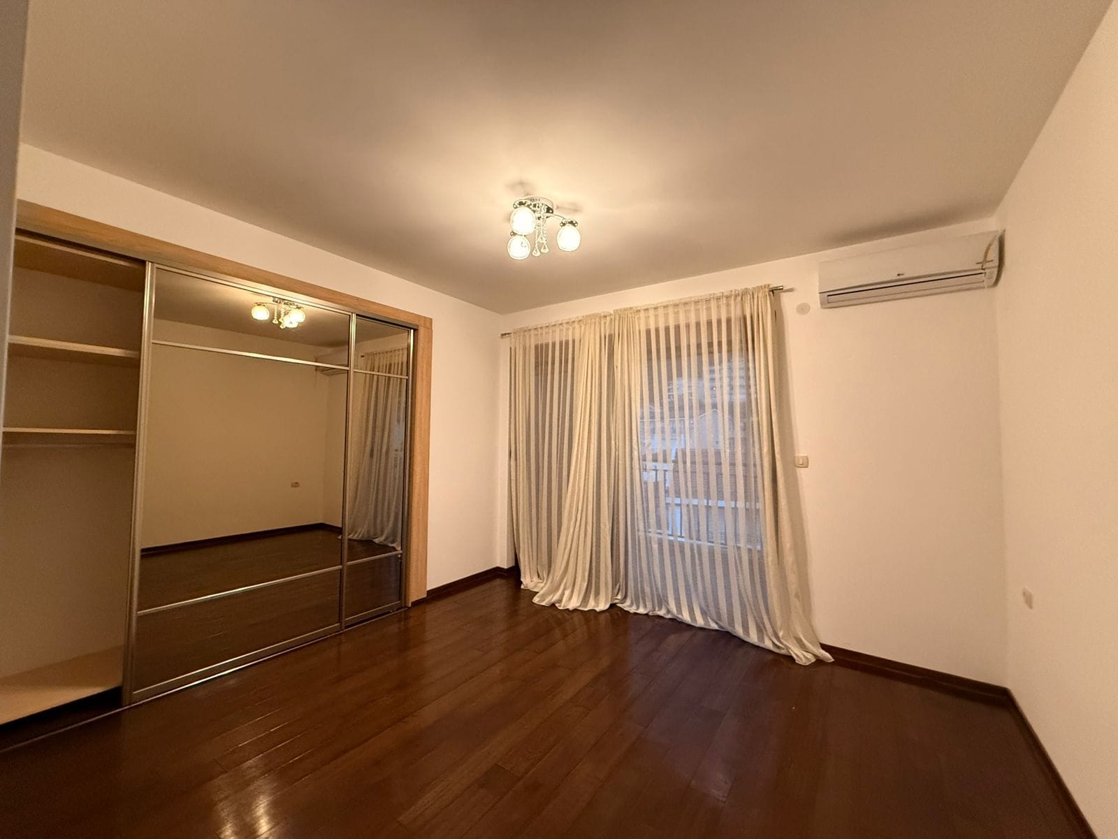 Bedroom in an apartment in Petrovac, Montenegro: mirrored sliding wardrobe, dark parquet flooring, light curtains, and air conditioning.