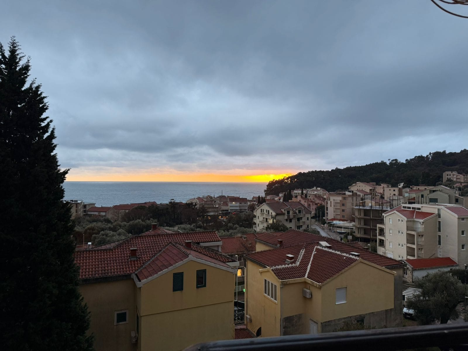 Panoramic view of the sea and the city of Petrovac in Montenegro at sunset, seen from an apartment window with tiled rooftops.