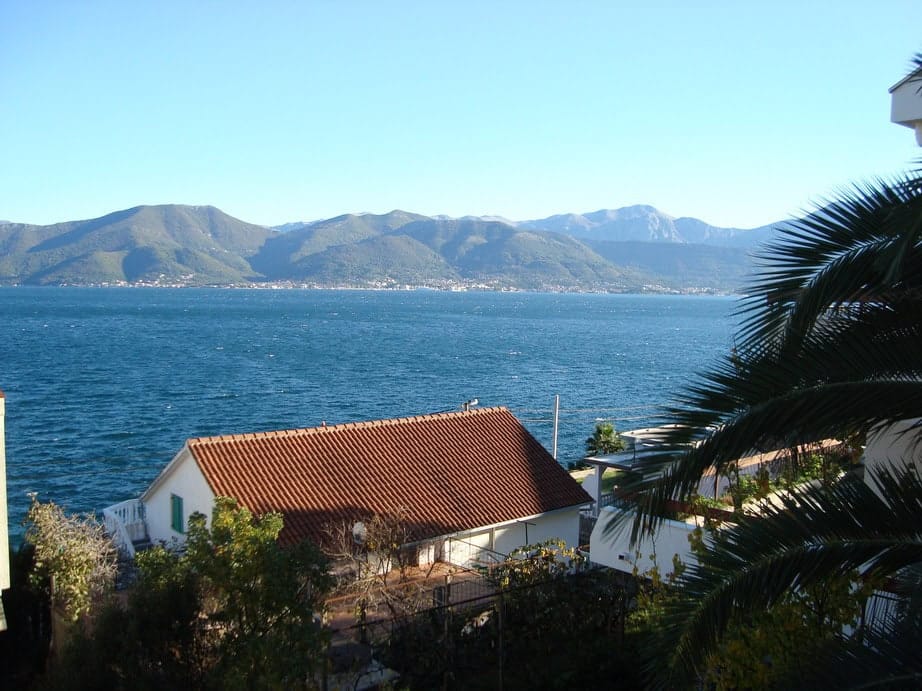Panoramic sea and mountain view from a house in Krašići, Montenegro: terrace, palm trees, and a house with a tiled roof by the water.