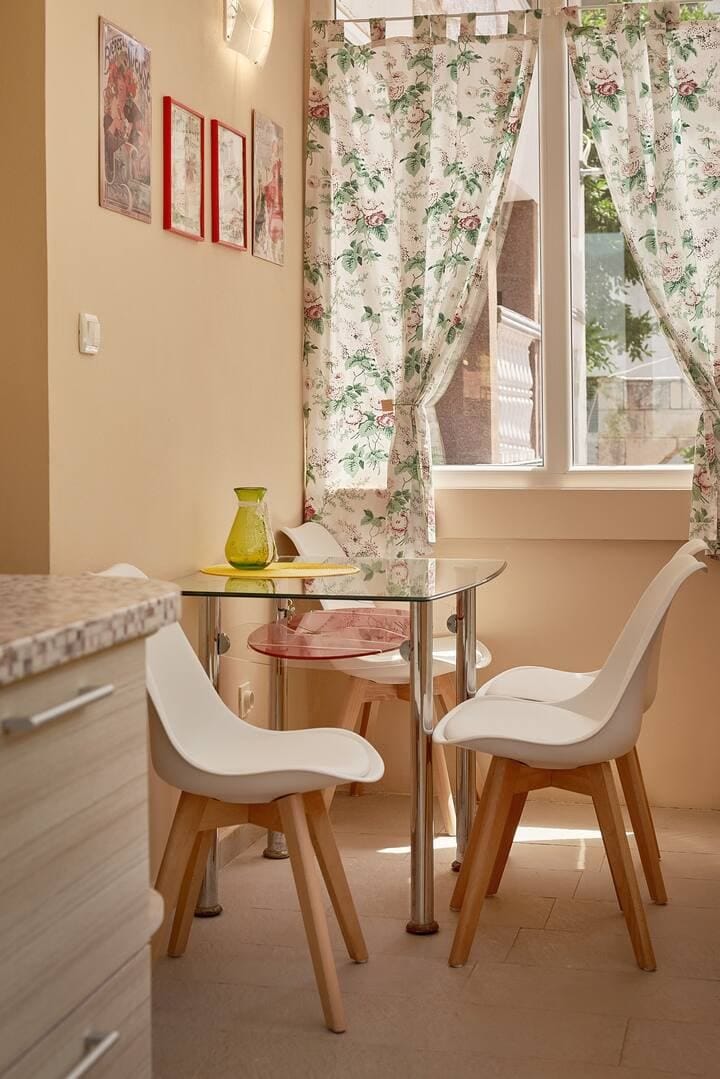 Cozy dining area in the kitchen of an apartment in Prčanj, Montenegro: a glass table and white chairs by the window with curtains.