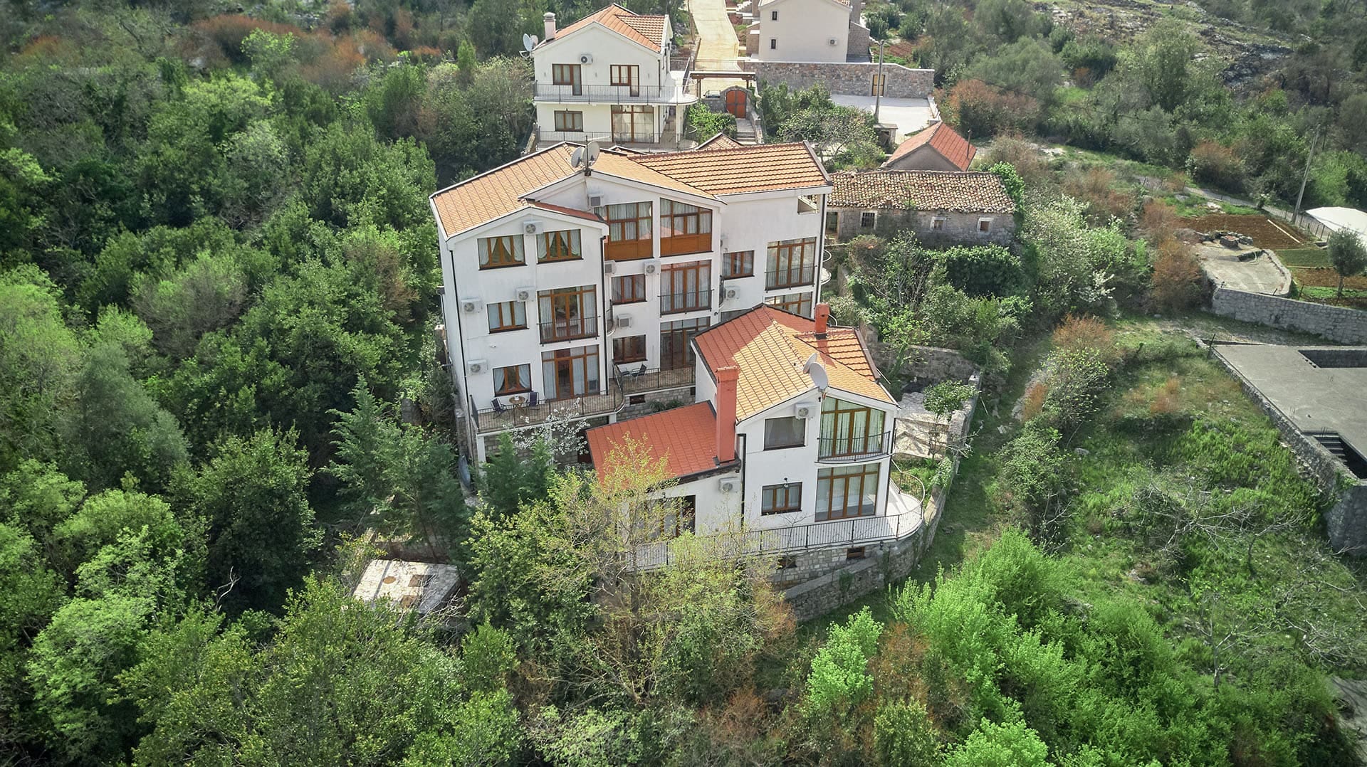 Aerial view of a townhouse in Vranovici, Bigovo, Montenegro: a white building with a tiled roof surrounded by lush greenery.