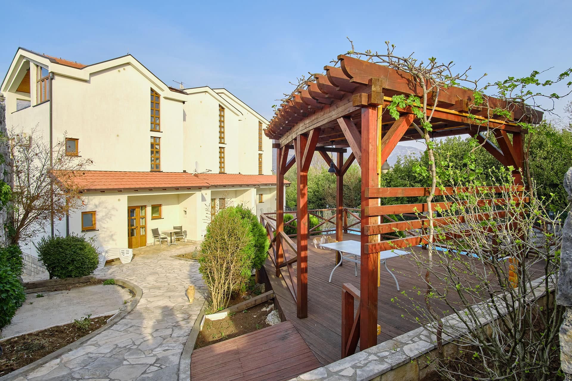 Townhouse courtyard in Bigovo, Montenegro: a wooden pergola, a terrace with furniture, and a stone path by the house.