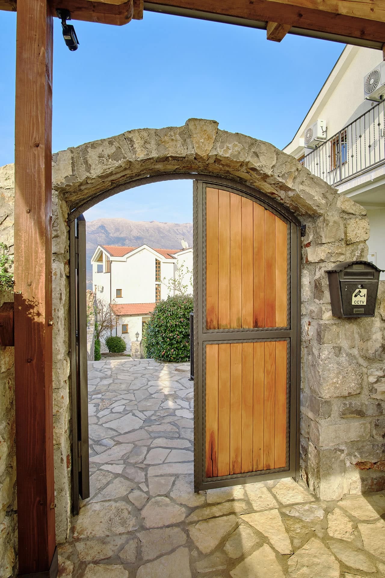 Stone arch and wooden gate by a house in Vranovici, Bigovo, Montenegro, with a view of the mountains and inner courtyard.