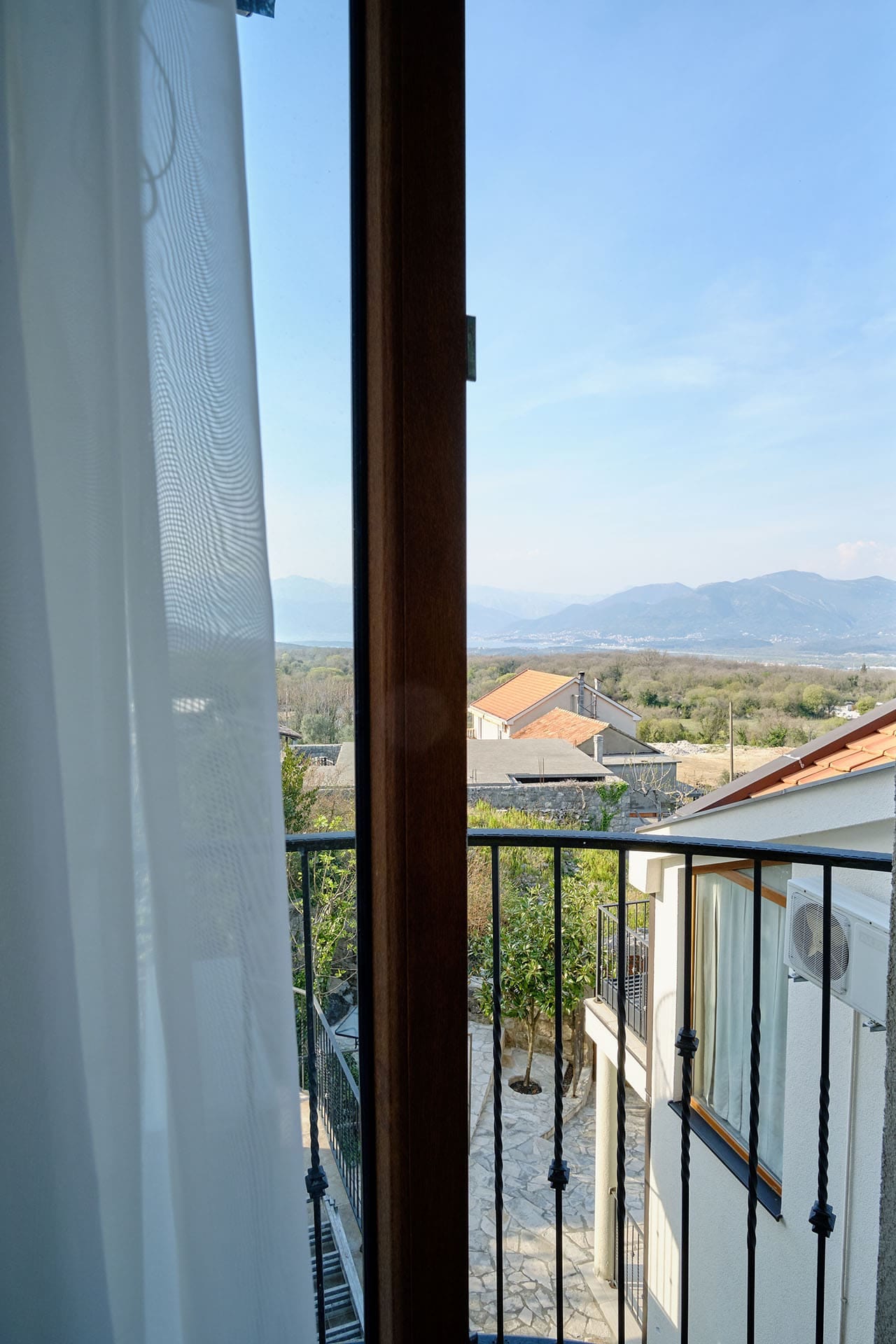 View from the townhouse balcony in Vranovici, Bigovo, Montenegro: a panorama of mountains, tiled roofs, and a green garden.