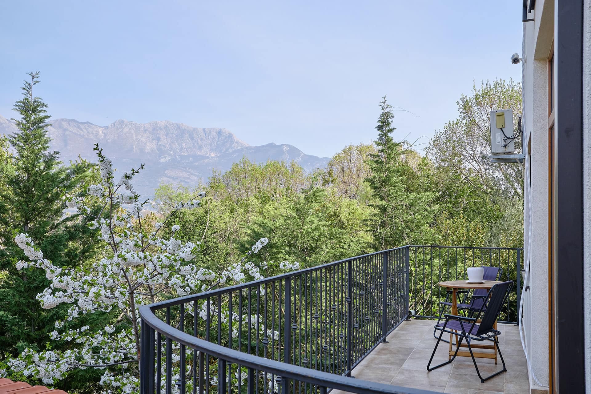 Townhouse balcony in Vranovici, Bigovo, Montenegro with a view of a blooming garden and mountains, table and chairs on the terrace.