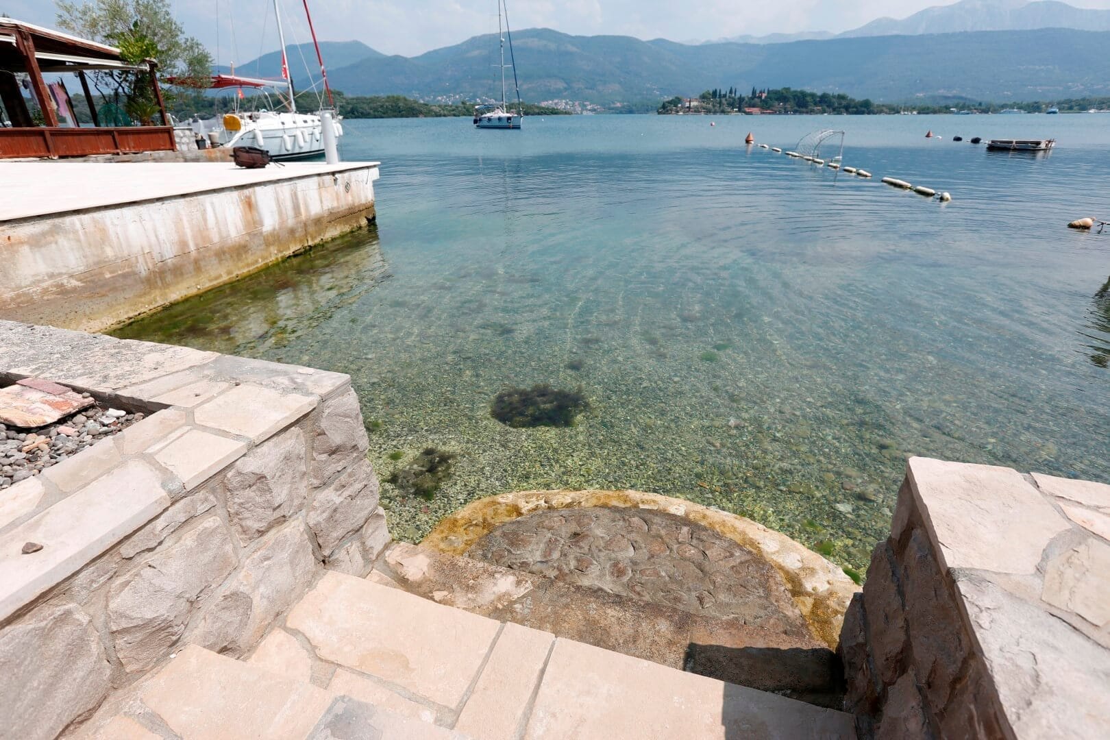Private beach and stone pier by a villa in Krašići, Montenegro: view of the clear sea, mountains, and yachts in the bay.
