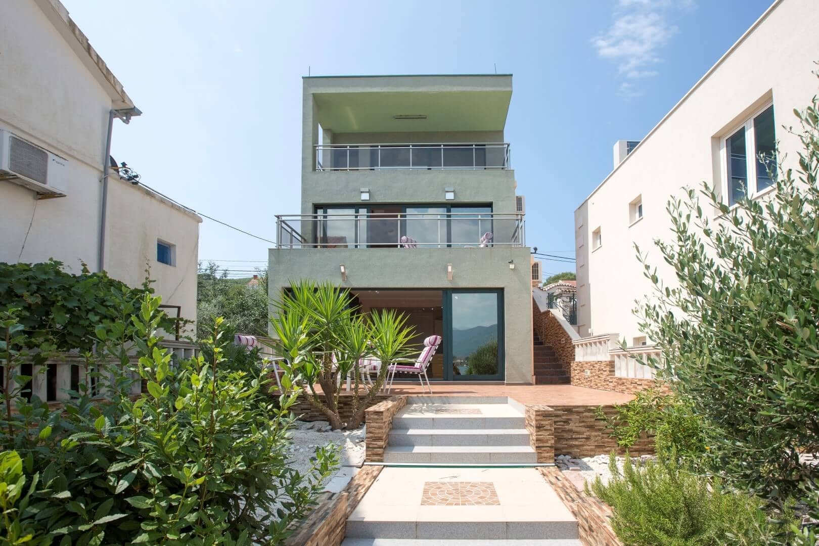 Three-story villa in Krašići, Montenegro: modern facade, terraces with panoramic glazing, and a cozy garden.