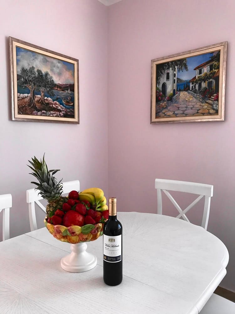 White dining table in the apartment living room in Dobri Vody, Montenegro, with a vase of fruit and wine in front of paintings.