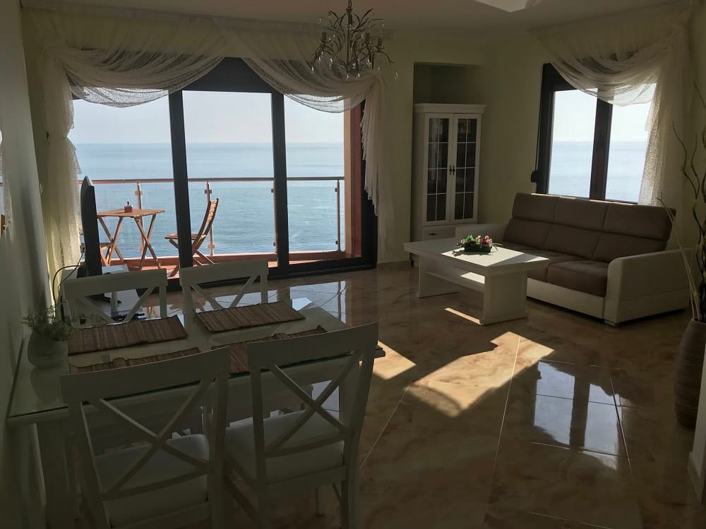 Living room with panoramic sea view in an apartment, Dobri Vody, Montenegro: light-colored furniture, terrace, and tiled floor.