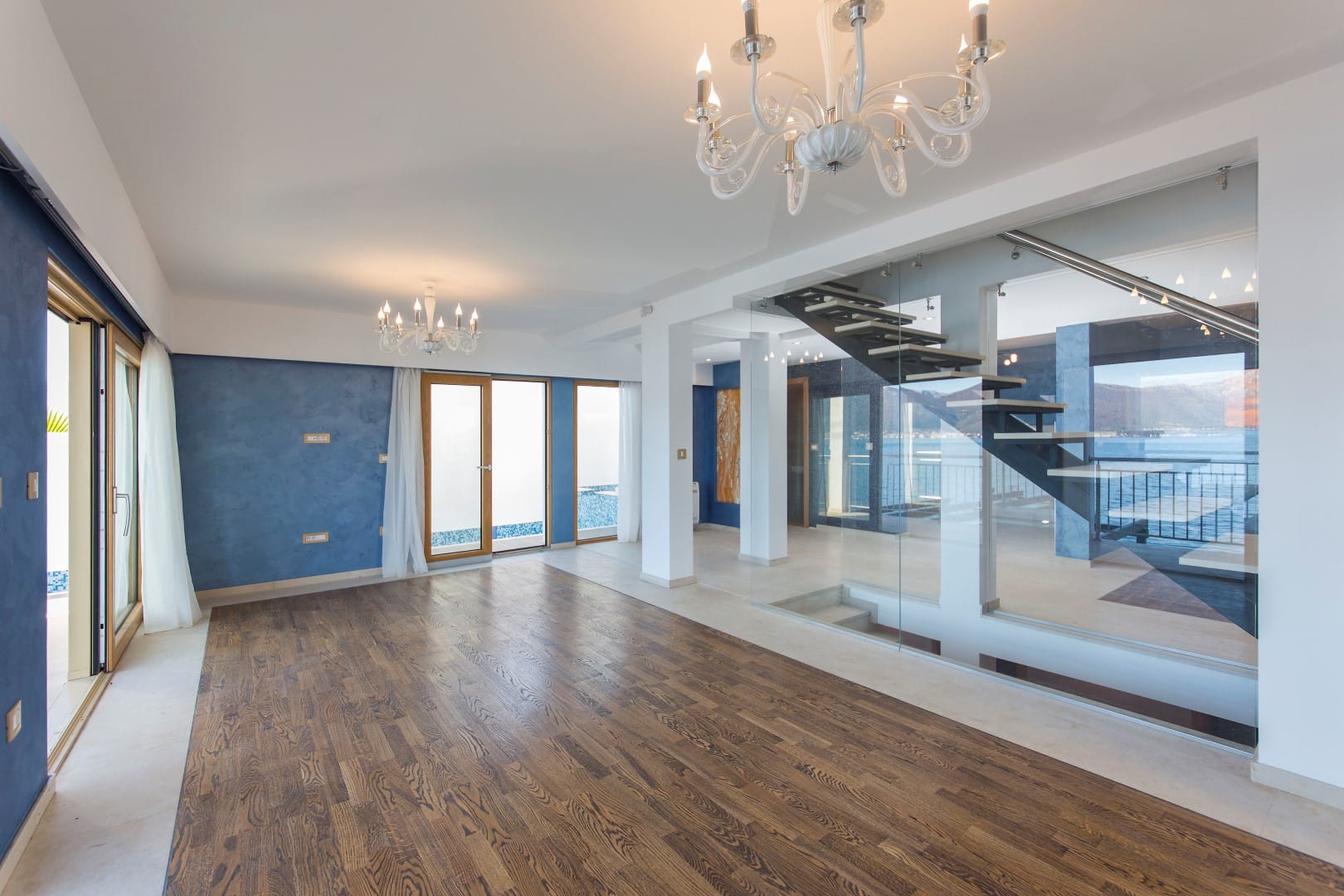 Spacious villa living room in Krašići, Montenegro: wooden floor, glass staircase, and panoramic sea view.