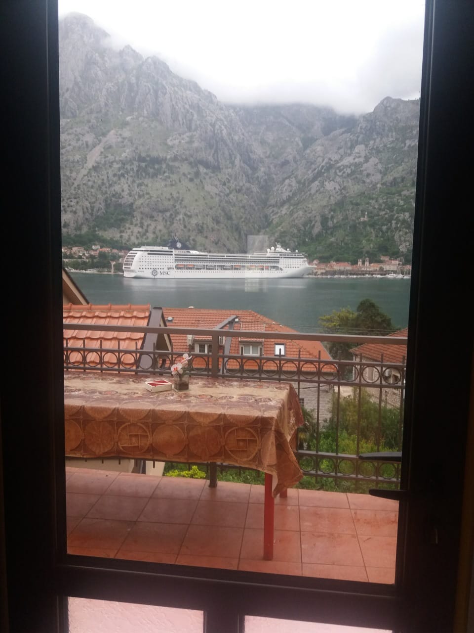 View from a house balcony in Muo, Montenegro: terrace with a table, panoramic view of the Bay of Kotor, mountains, and a cruise ship.