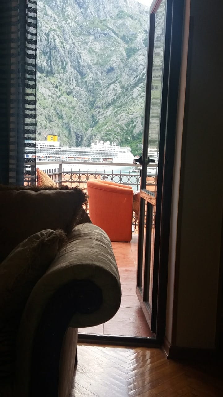 View from the living room of a house in Muo, Montenegro: a terrace with an orange chair and a panoramic view of the mountains and a cruise ship.