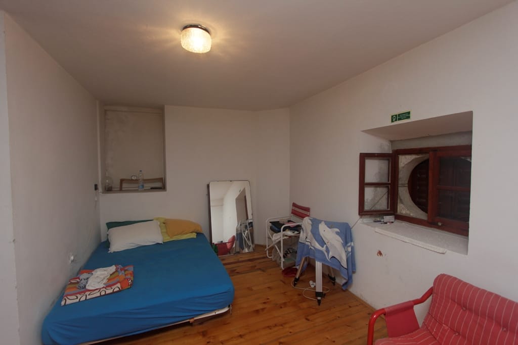 Bedroom in a house in Prčanj, Montenegro: a room with a wooden floor, bed, mirror, and an antique window in a niche.