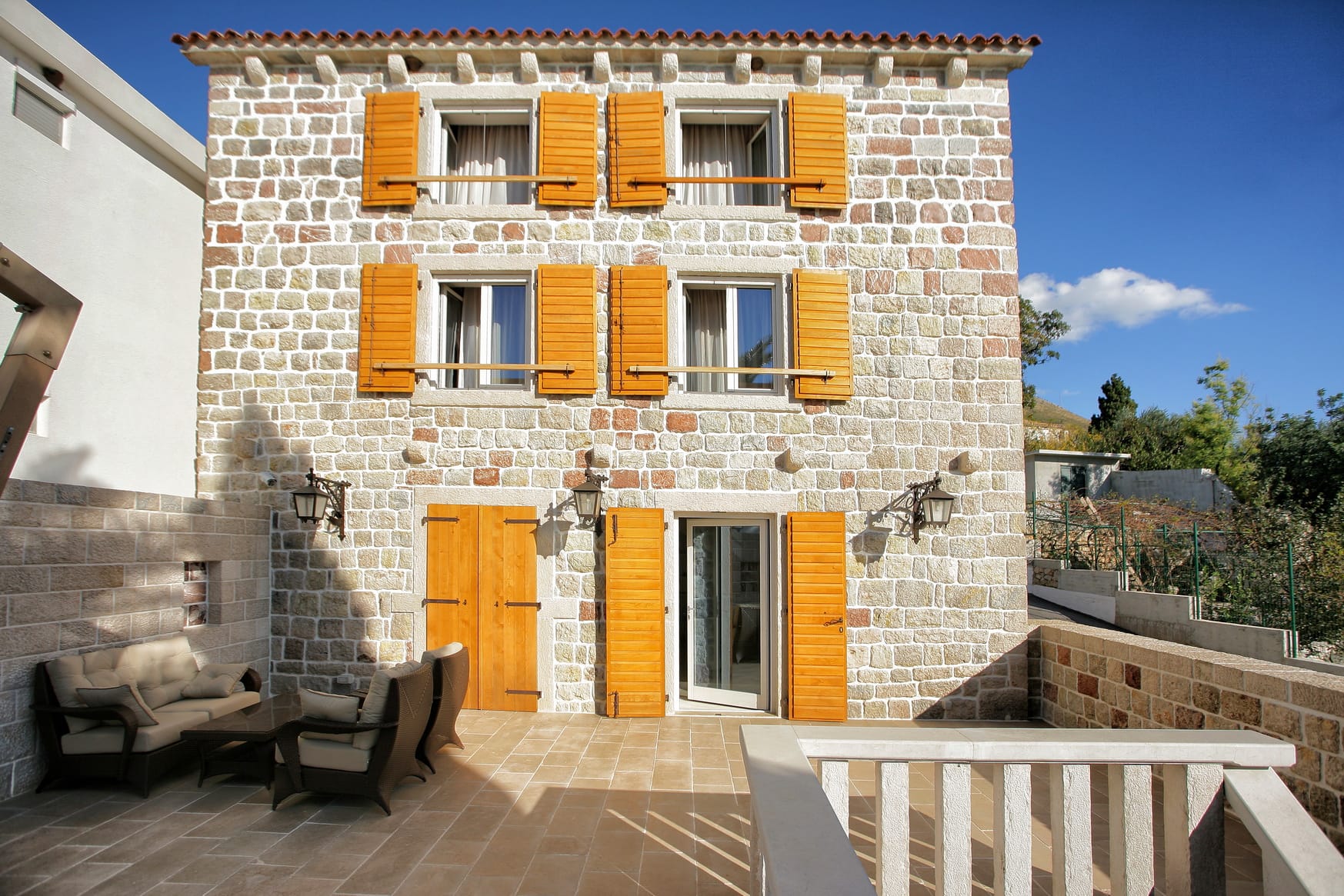 Three-story stone villa in Reževići, Montenegro, with a terrace, wicker furniture, and wooden shutters on the windows.