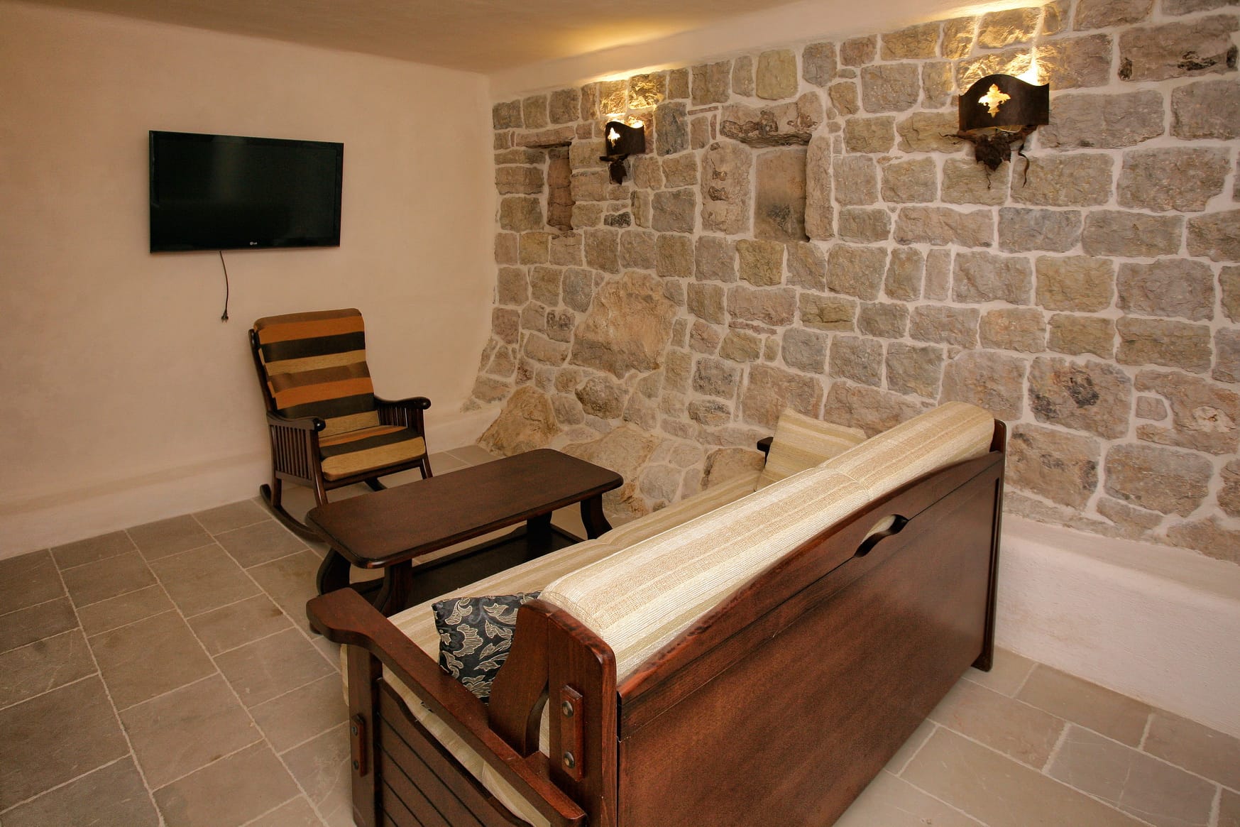 Living room with a stone wall and wooden furniture in a villa in Reževići, Montenegro. Cozy interior with a TV and an armchair.
