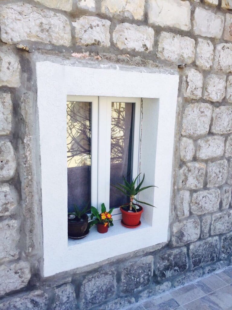 White plastic window in the stone wall of a mini-hotel in Rafailovici, Montenegro, with potted plants on the windowsill.