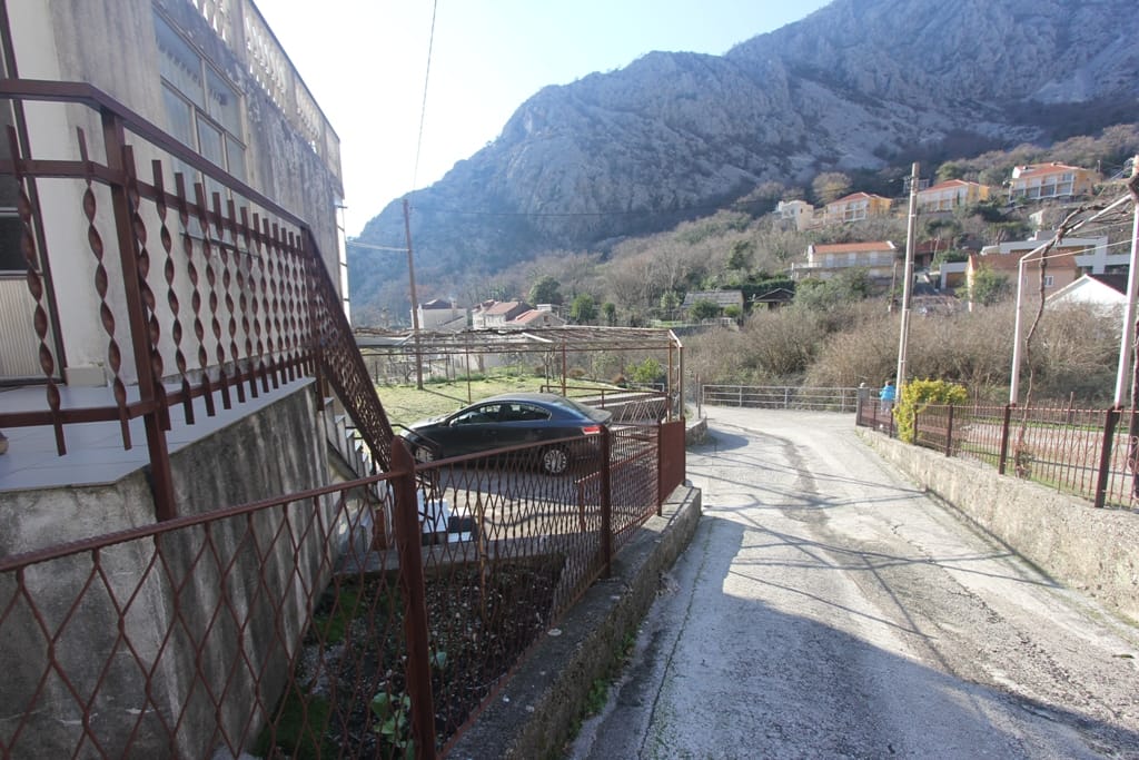 View of a house in Orahovac, Montenegro: the surrounding area with parking, a metal fence, and a mountain landscape.