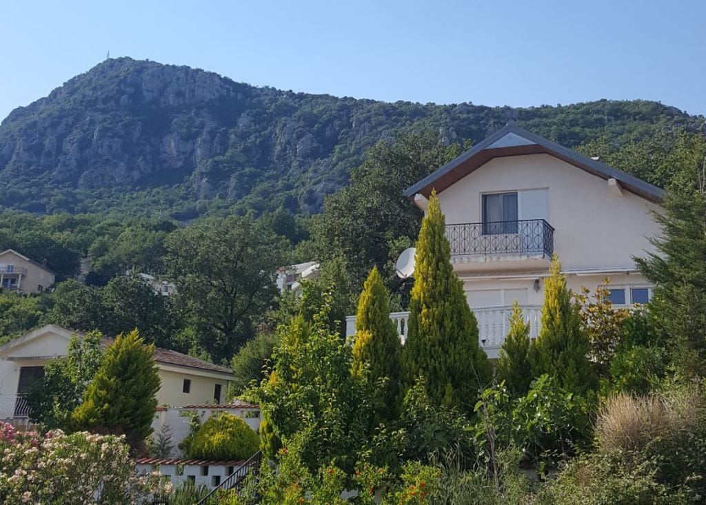 Two-story house with a balcony surrounded by mountains and greenery, Sutomore, Montenegro. Front view of a private house with a garden.