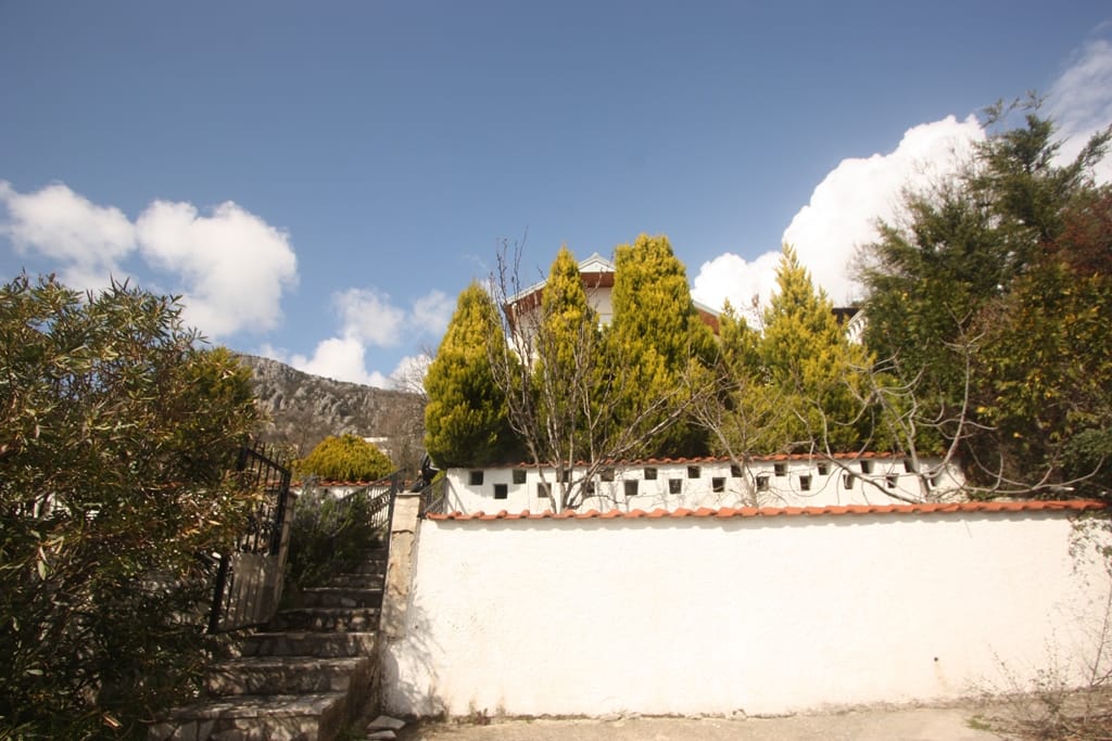 White house with a tiled roof and stone staircase in Sutomore, Montenegro, surrounded by cypress trees and mountain scenery.