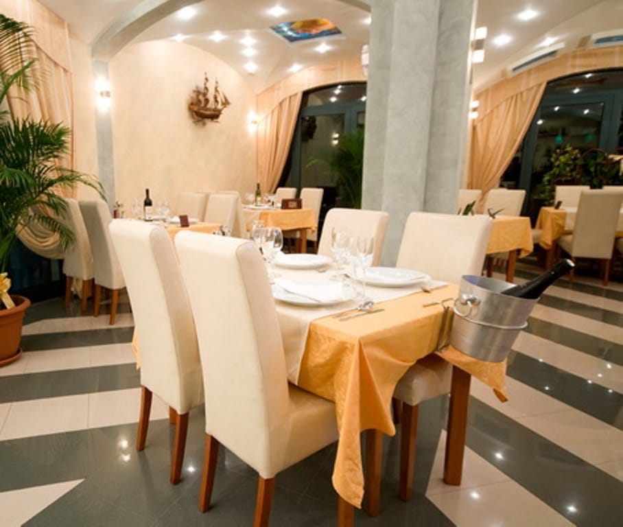 Restaurant interior in a mini-hotel in Bijela, Montenegro: set tables, light-colored furniture, and tiled flooring in the dining room.