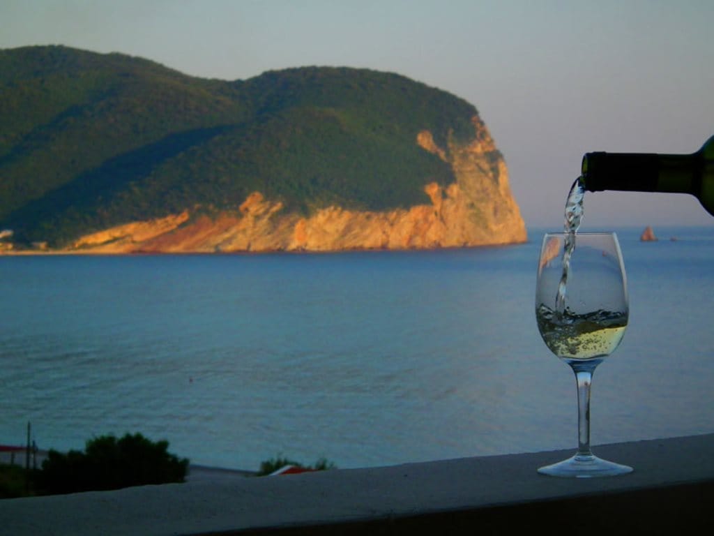 View from a hotel balcony in Buljarica, Montenegro: a glass of wine against the seaside coast and scenic cliffs at sunset.