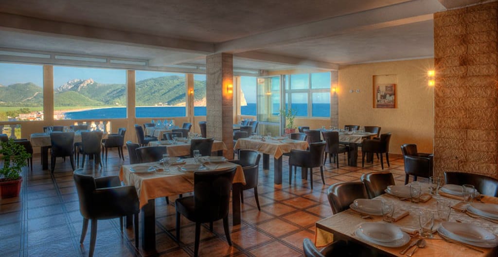 Hotel restaurant interior in Buljarica, Montenegro, with panoramic views of the sea and mountains and set tables.