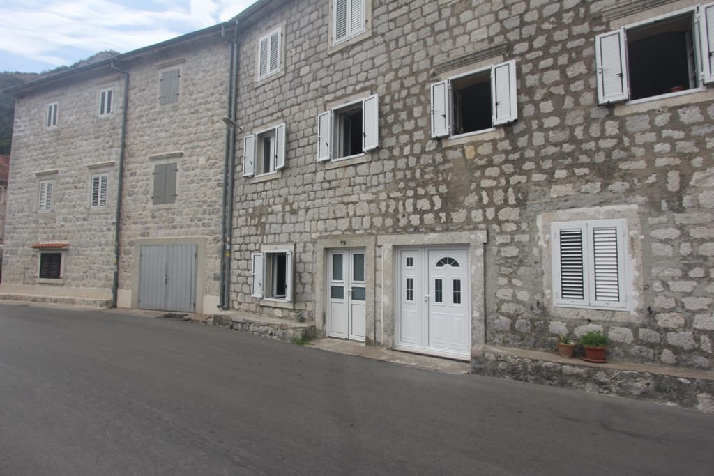 Facade of a traditional stone house in Muo, Montenegro, with white doors and window shutters along the road.