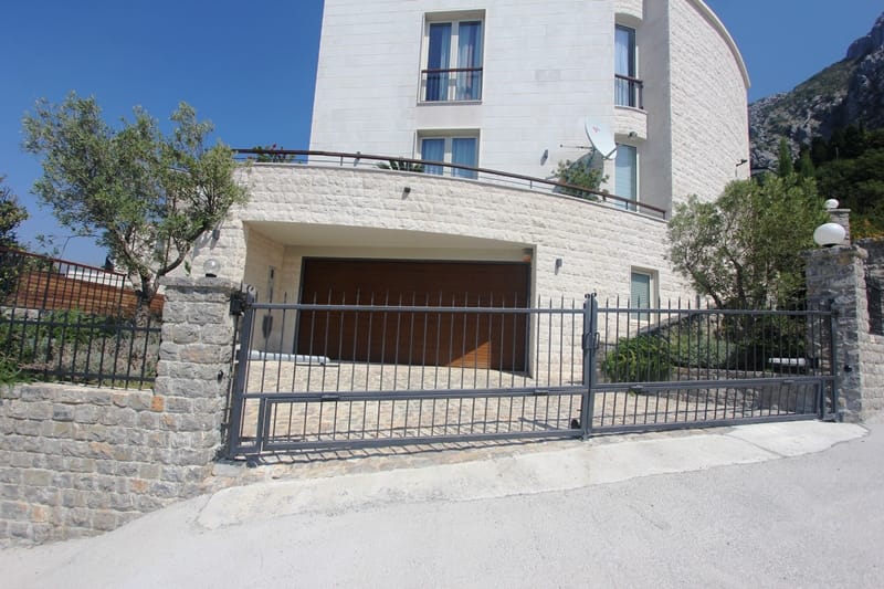 Modern villa in Blizikuće, Montenegro: light stone facade, garage, and wrought-iron gates against a mountain backdrop.