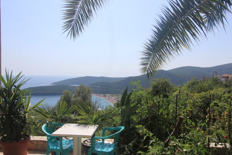 View from the house terrace in Seoce, Montenegro: a table with plastic chairs, palm trees, and a panoramic view of the sea and mountains.