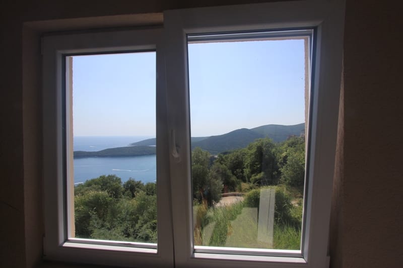 View from a plastic window of the sea and mountains in a house in Seoce, Montenegro: a panoramic landscape with greenery and coastline.