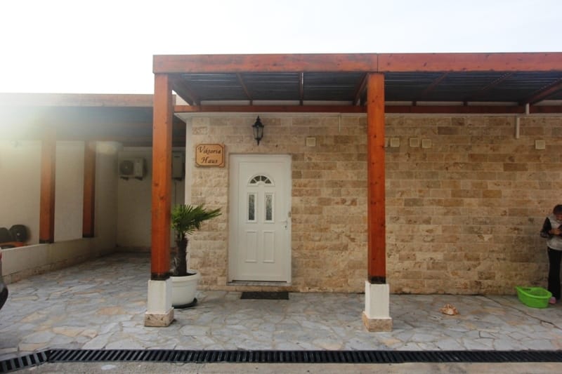 Facade of the Viktoria Haus building in Utjeha, Montenegro: stone cladding, a covered terrace with wooden supports, and the entrance door.