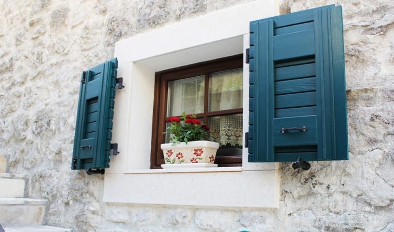 Window of a stone house in Strp, Montenegro, with green wooden shutters and a flower pot on the windowsill.