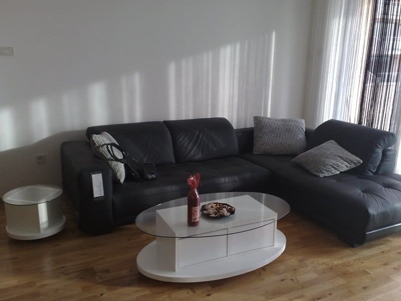 Living room in an apartment in Pržno, Montenegro: black leather corner sofa, white oval coffee table, and parquet flooring.