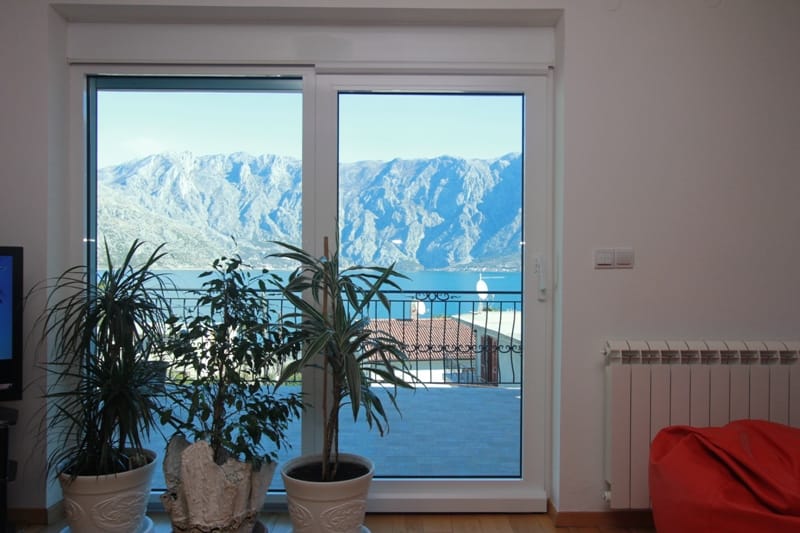 Sea and mountain view from the living room of a house in Stoliv, Montenegro: panoramic window, houseplants, and a bright interior.