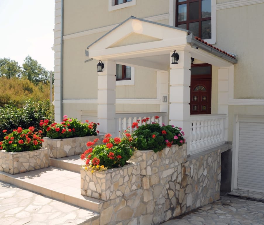 Entrance to a villa in Markovichi, Montenegro: a porch with stone cladding, white columns, and blooming geraniums.