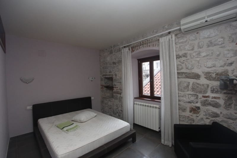 Bedroom with a stone wall and a double bed in a villa in Prčanj, Montenegro: view of tiled roofs from the window.