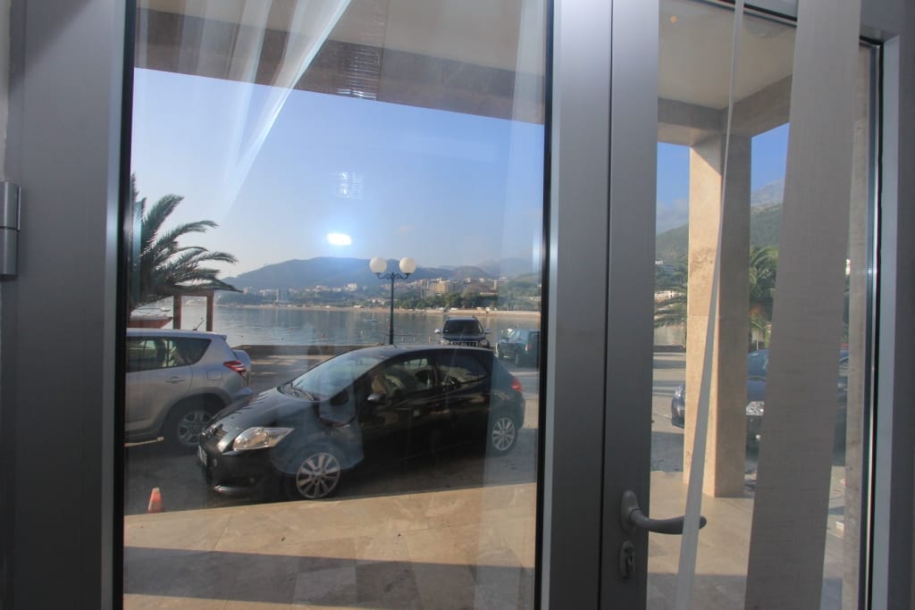 View from the window of a commercial space overlooking the sea, beach, and mountains in Rafailovici, Montenegro, with parking in front of the entrance.