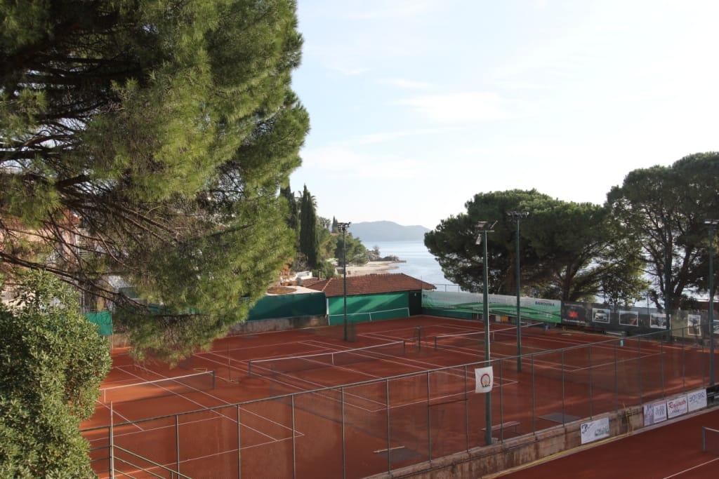 View of clay tennis courts and the seashore near a hotel in Igalo, Montenegro, surrounded by coniferous trees.