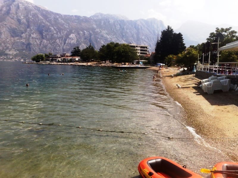 View of the beach and mountains from a plot of land in Stoliv, Montenegro: coastline, clear sea, and a pier.