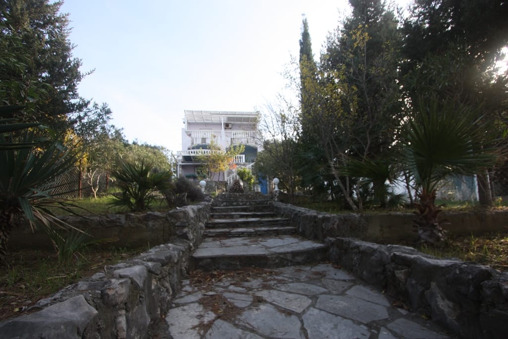 Three-story house in Utjeha, Montenegro, with a stone staircase, terraces, and a green garden on the adjacent grounds.