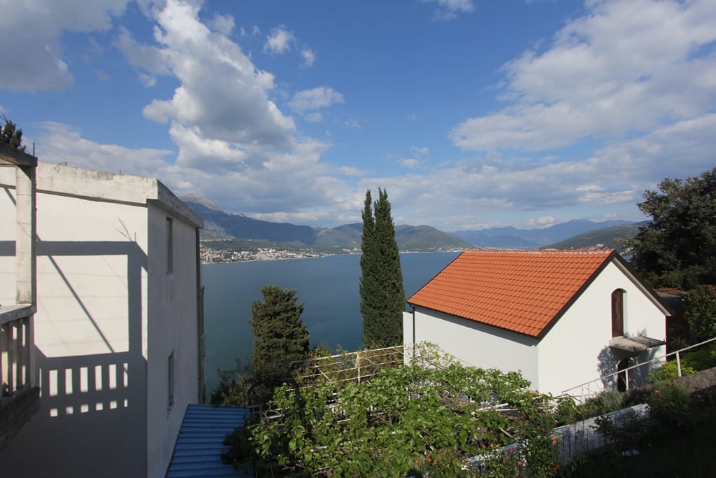 Panoramic view of the Bay of Kotor and mountains from a plot in Njivice, Igalo, Montenegro: houses with tiled roofs and greenery.