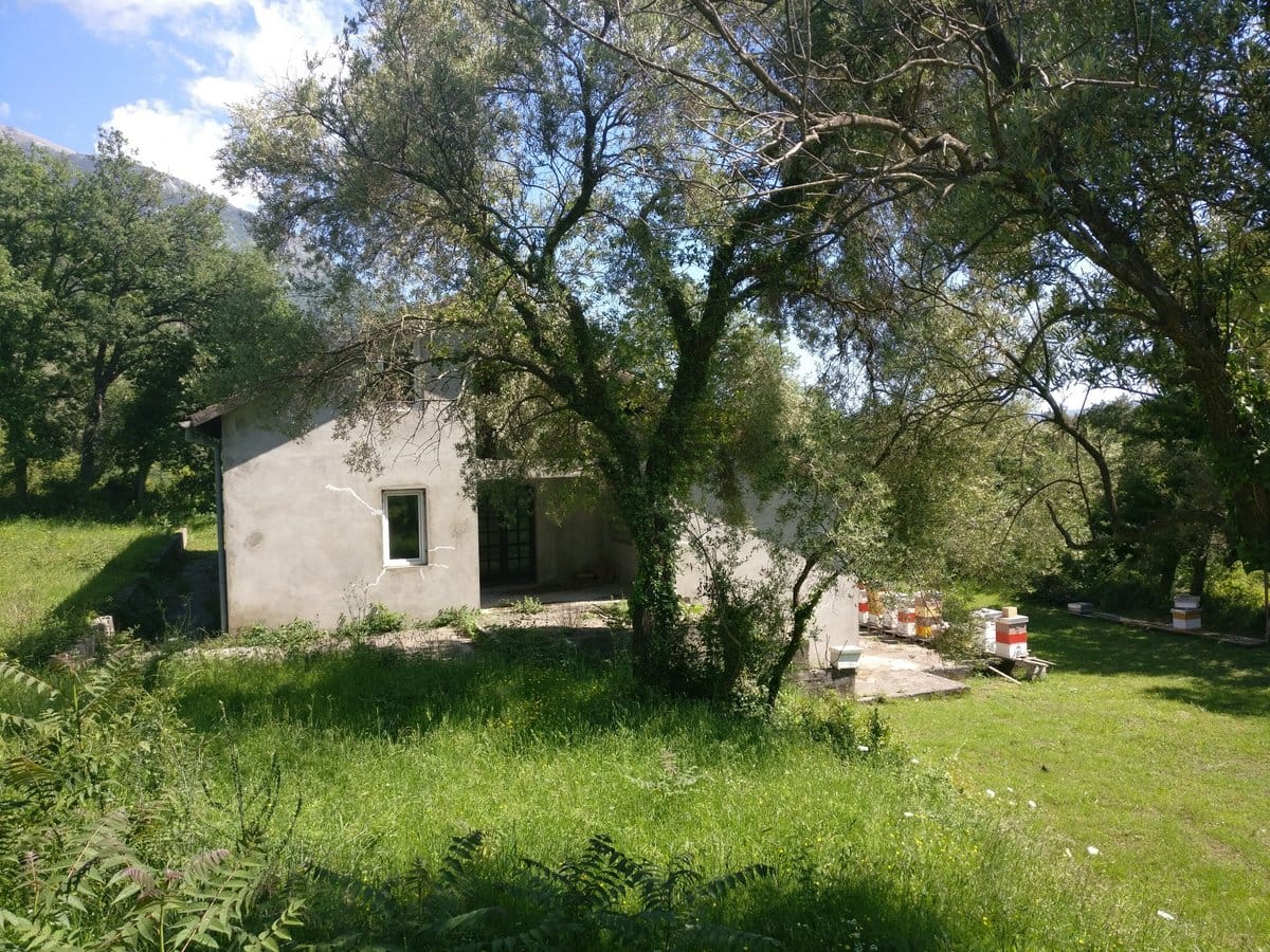 Two-story house in Kavac, Montenegro, with a plot, garden, and apiary against a backdrop of mountains and green trees.