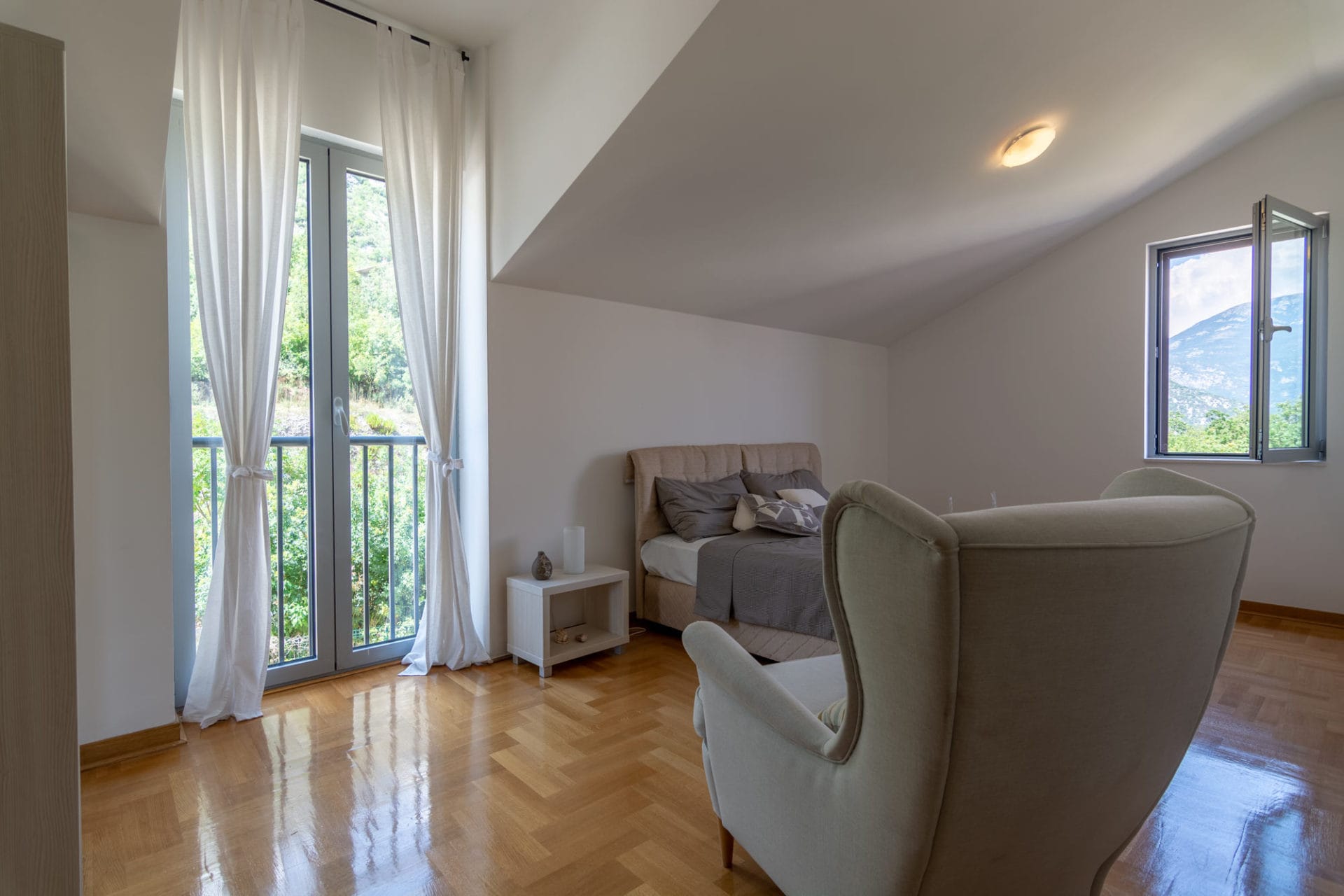 Bright bedroom in an apartment in Morinj, Montenegro: a double bed, an armchair, parquet flooring, and a mountain view from the window.