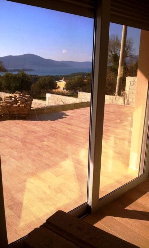 View from the living room onto the terrace and the sea in Kavac, Montenegro: panoramic windows, tiled floor, and mountain scenery.
