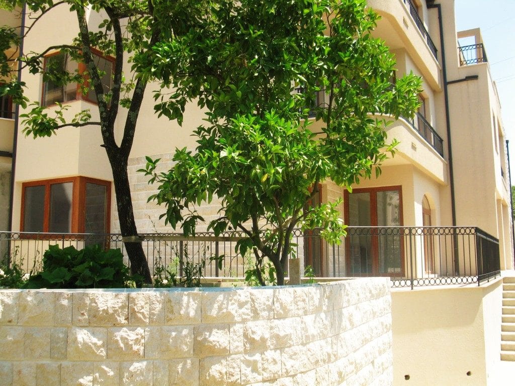 Facade of a residential complex in Igalo, Montenegro: stone cladding, terraces with wrought-iron railings, and a green tree in the courtyard.